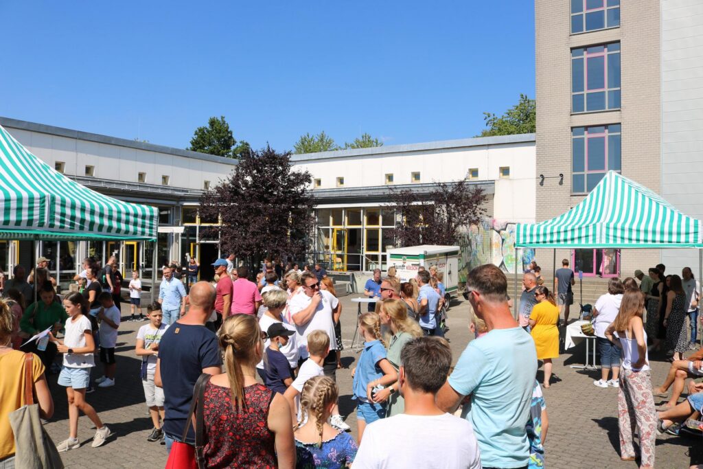 Vielerlei Menschen stehen auf einem Schulhof bei Ständen mit grünen Markisen.