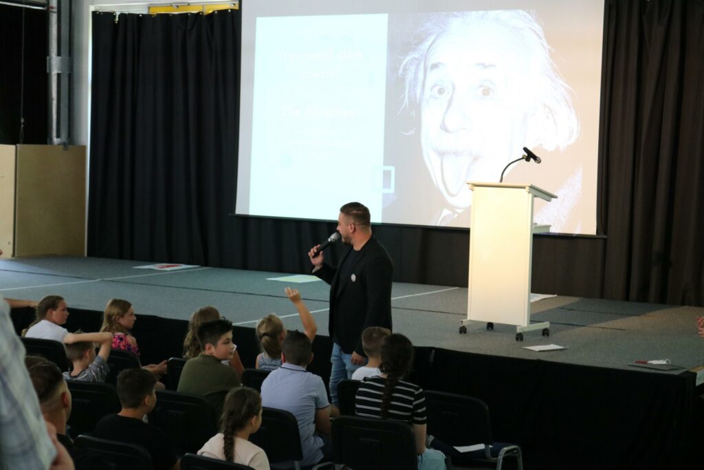 Vortragender spricht vor einer Gruppe von Schülern, im Hintergrund ein Bild von Einstein.