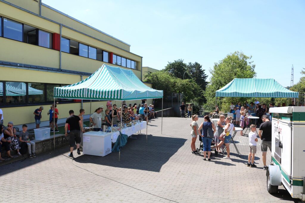 Menschen stehen an Ständen auf einem Schulhof mit Zelten an sonnigem Tag.