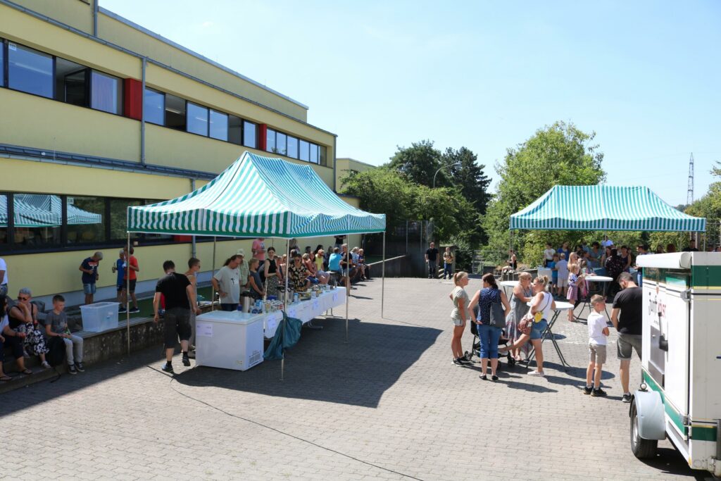Schulhof mit Ständen und Besuchern bei schönem Wetter.