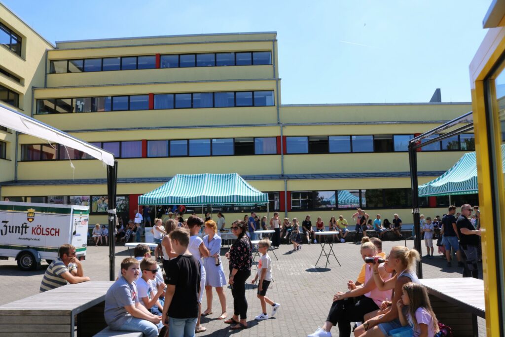 Schüler stehen und sitzen auf dem Schulhof vor einem Gebäude. Im Hintergrund sind Stände.