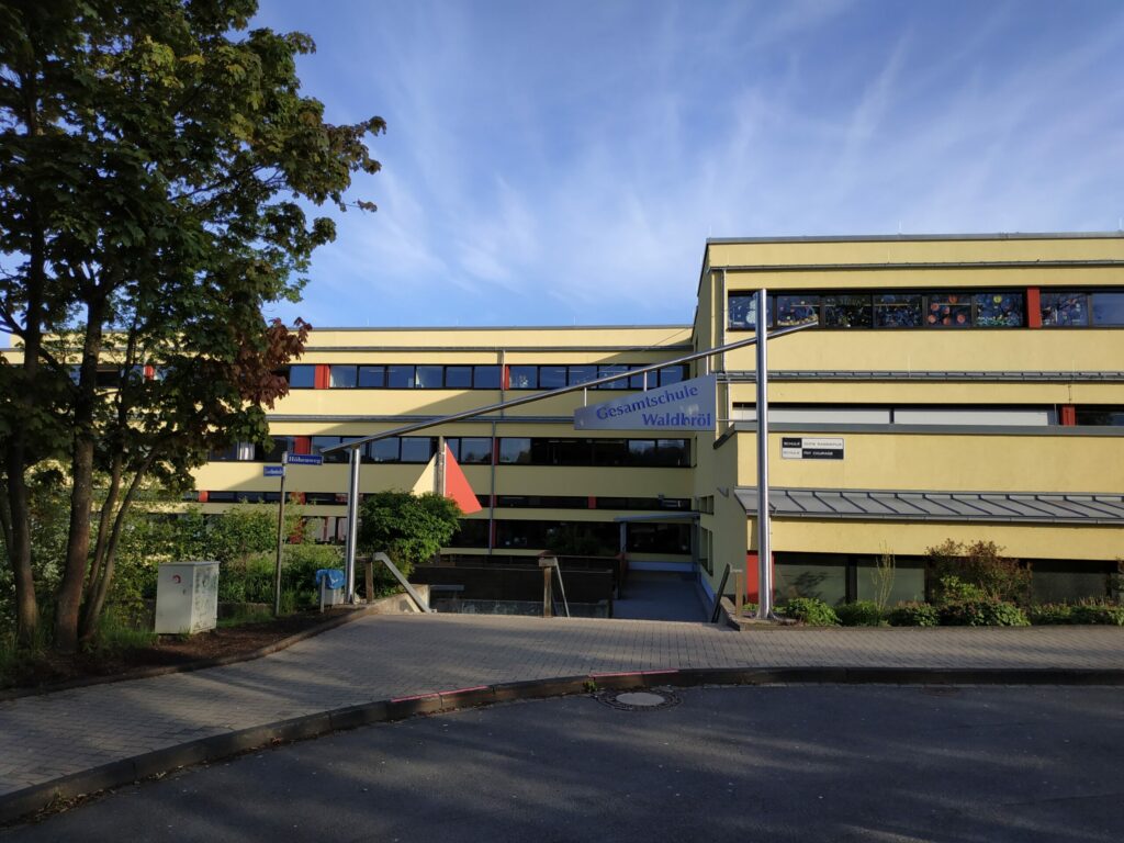 Schulgebäude mit gelber Fassade und Flaggen im Vordergrund.