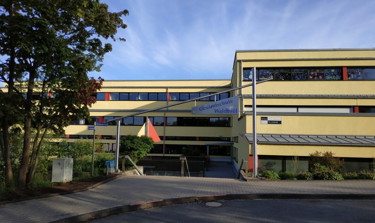 Schulgebäude mit gelber Fassade und Flaggen im Vordergrund.