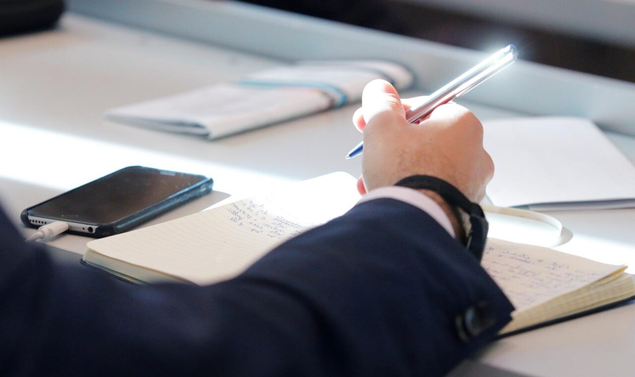 Eine Hand hält einen Stift über einem Notizbuch auf einem Tisch.