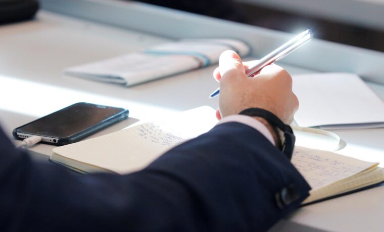 Eine Hand hält einen Stift über einem Notizbuch auf einem Tisch.