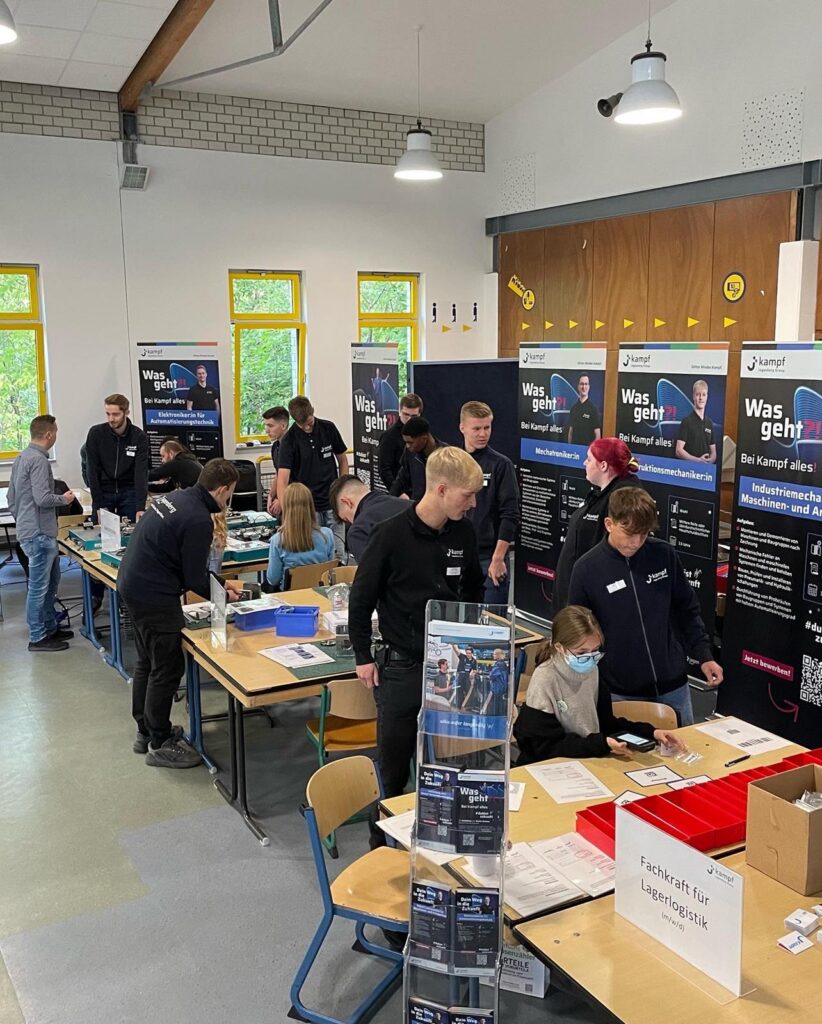 Schüler arbeiten an Tischen in einem Klassenraum, Informationsstände im Hintergrund.