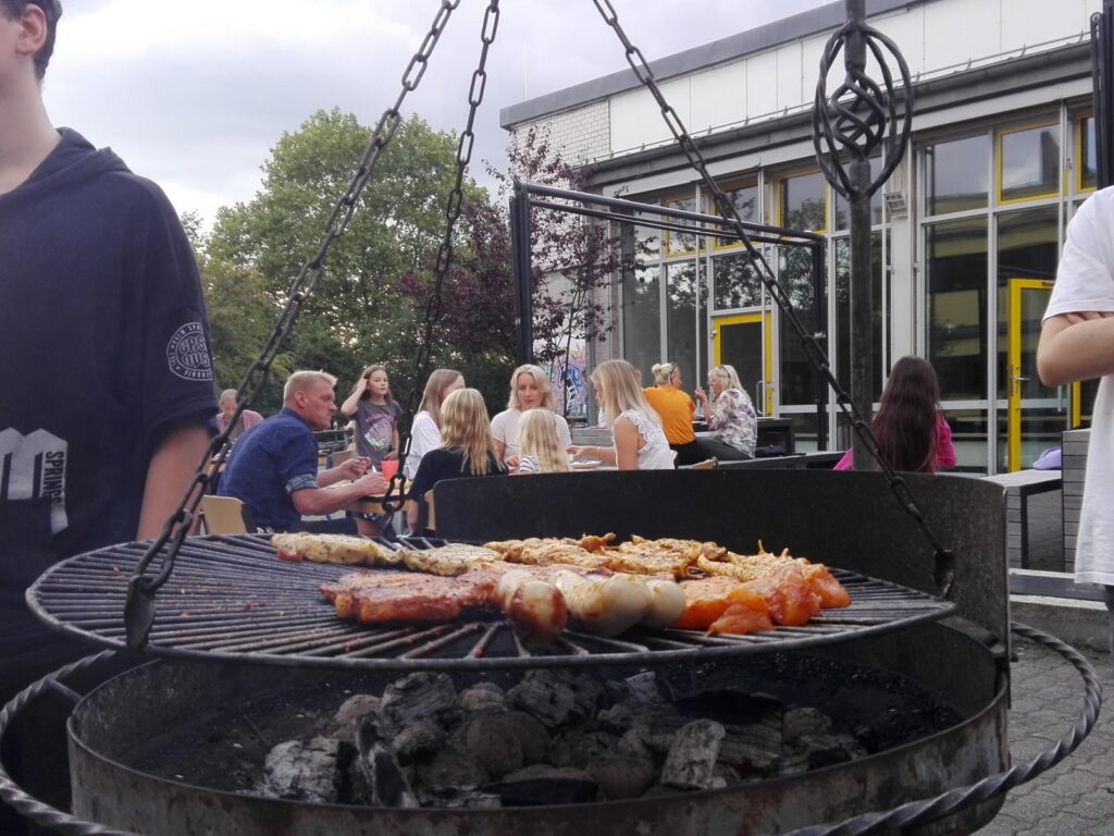 Grill mit verschiedenen Fleischsorten, im Hintergrund Schülerinnen und Schüler.