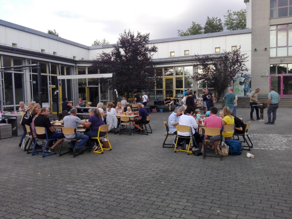 Schüler und Lehrer sitzen an Tischen im Schulhof und unterhalten sich.