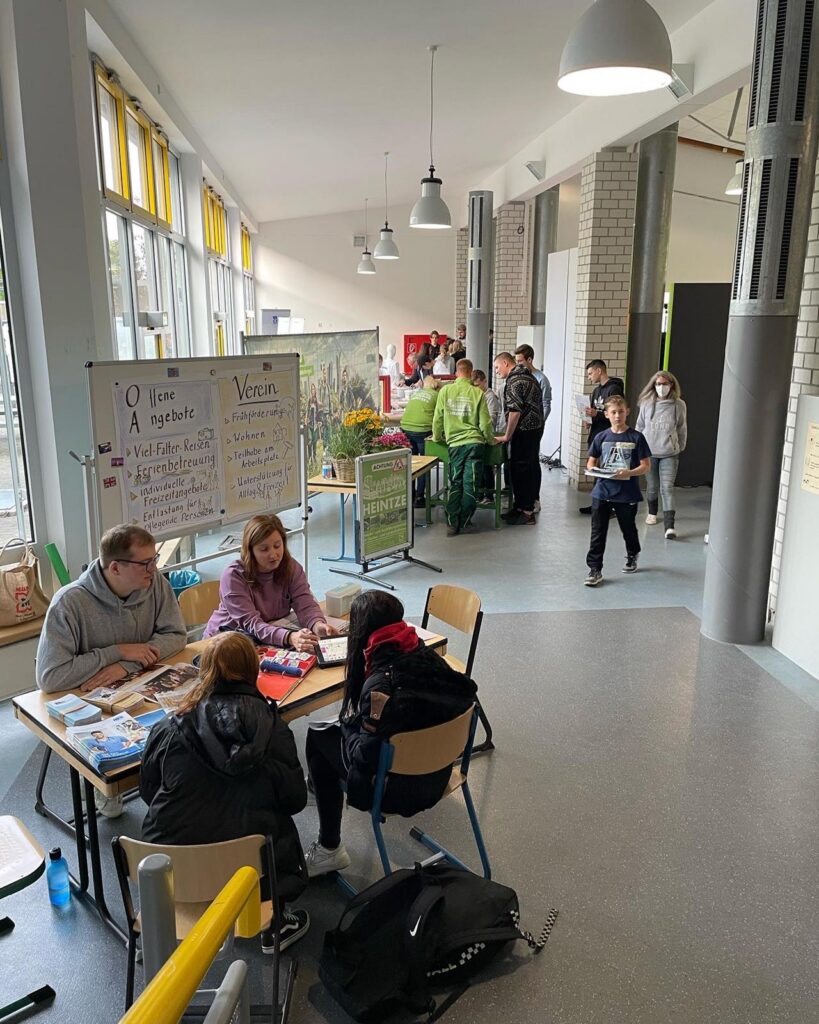 Schüler arbeiten an Tischen, andere stehen in einem Flur. Plakate sind sichtbar.
