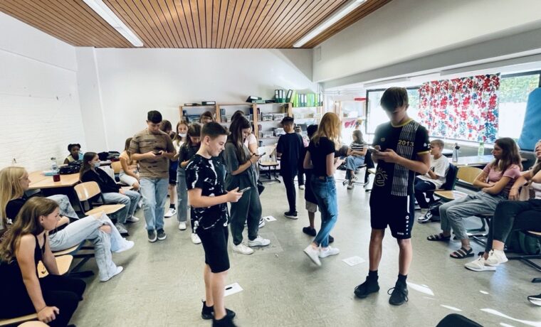 Schülerinnen und Schüler stehen in einem Klassenraum und interagieren miteinander.