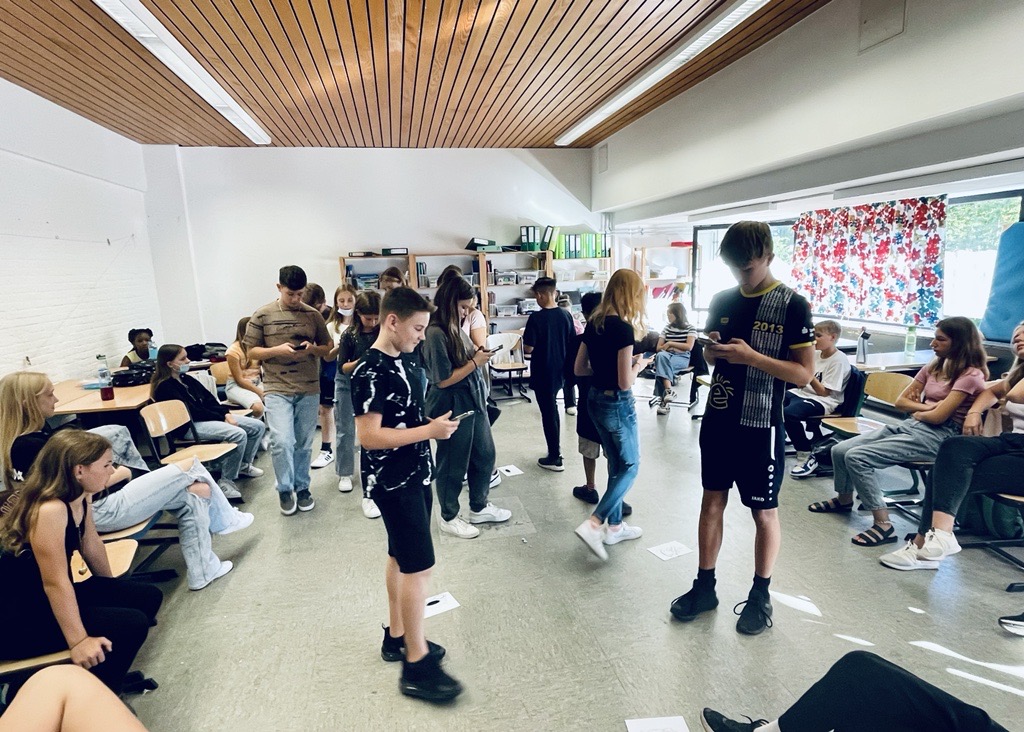 Schülerinnen und Schüler stehen in einem Klassenraum und interagieren miteinander.