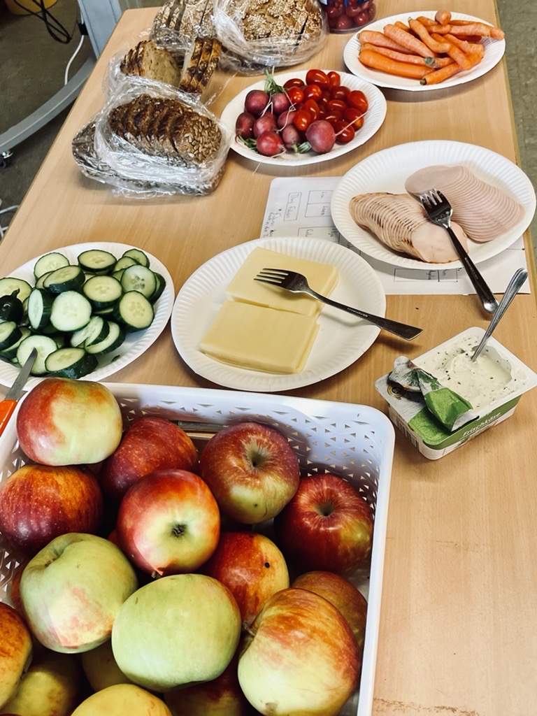 Tisch mit verschiedenen Lebensmitteln: Äpfel, Gemüse, Aufschnitt und Brot.
