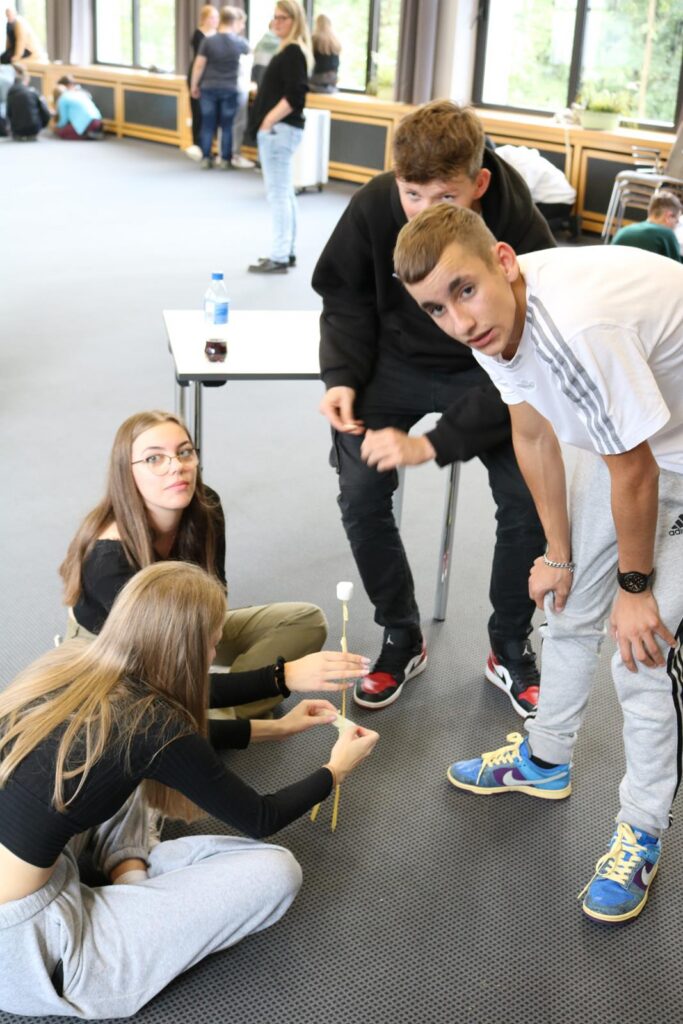 Schüler arbeiten gemeinsam an einem Tisch, einige sitzen, andere stehen.
