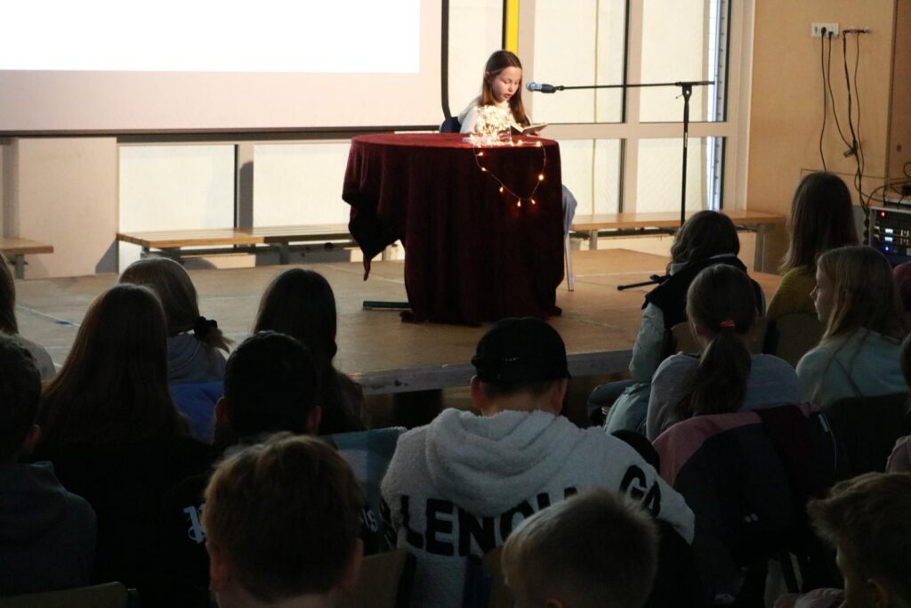 Schülerin liest am Tisch vor Publikum in einem Klassenraum.