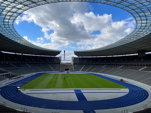 Blick auf ein modernes Stadion mit Rasenfläche und Laufbahn unter einem wolkigen Himmel.