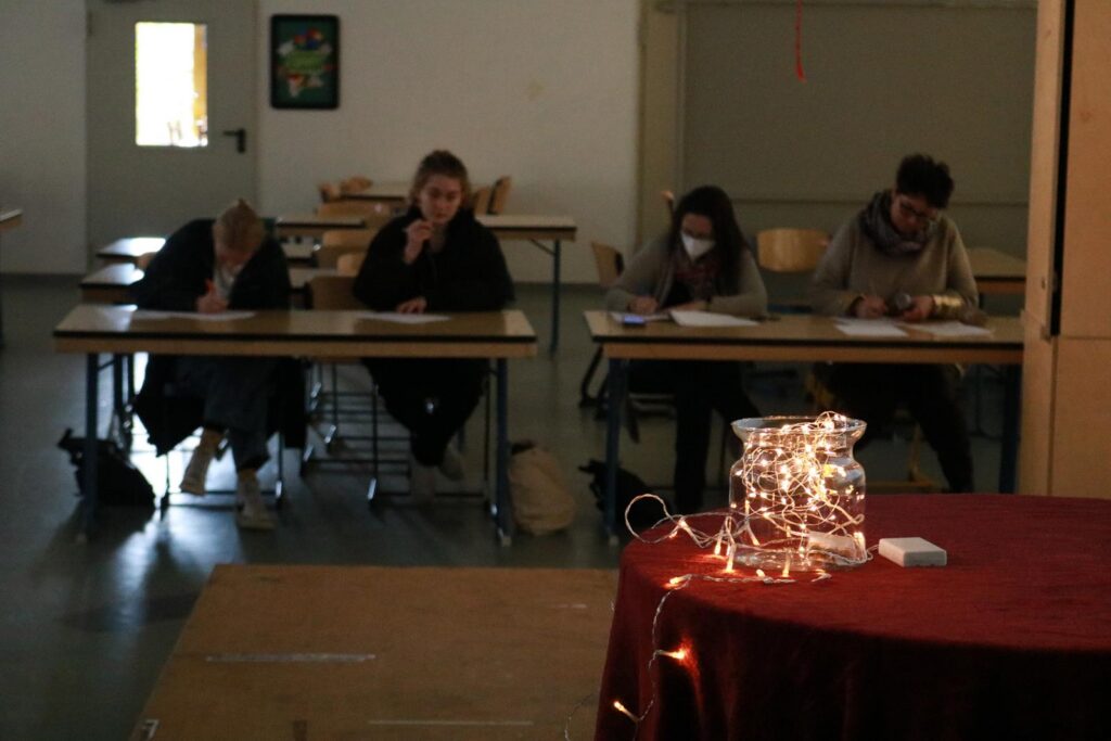 Schülerinnen und Schüler sitzen an Tischen und schreiben. Im Vordergrund steht ein Tisch mit Lichtern.