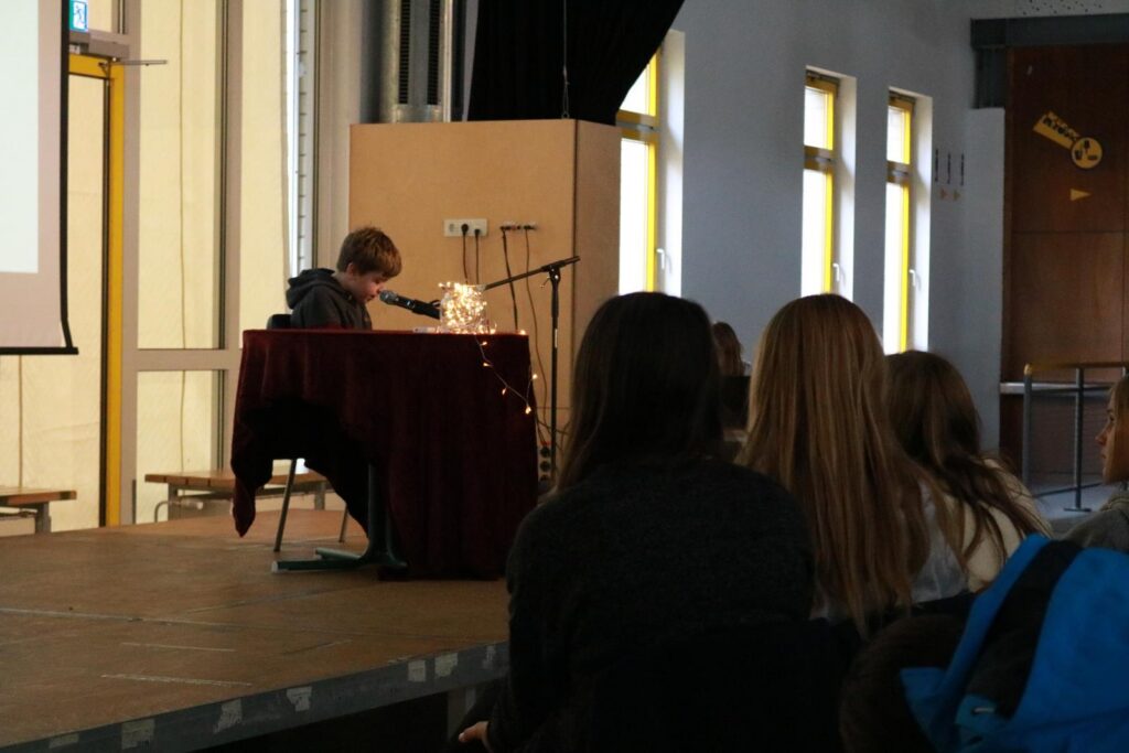 Ein Schüler sitzt an einem Tisch und präsentiert vor Publikum.