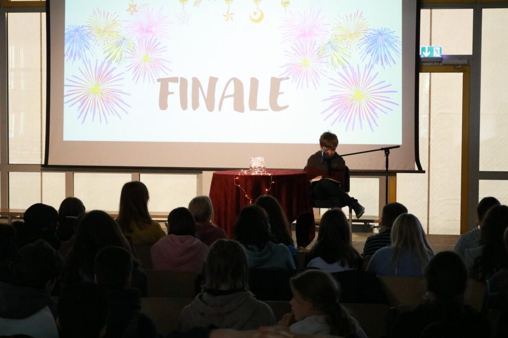 Person sitzt auf der Bühne vor einem Publikum, Hintergrund mit "FINALE" und bunten Mustern.