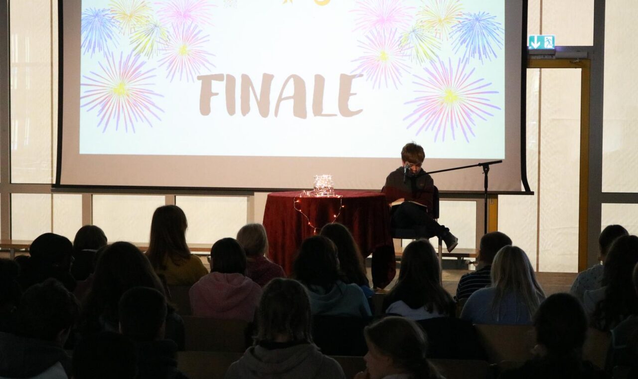 Person sitzt auf der Bühne vor einem Publikum, Hintergrund mit "FINALE" und bunten Mustern.