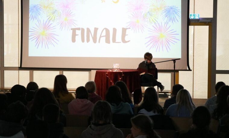 Person sitzt auf der Bühne vor einem Publikum, Hintergrund mit "FINALE" und bunten Mustern.