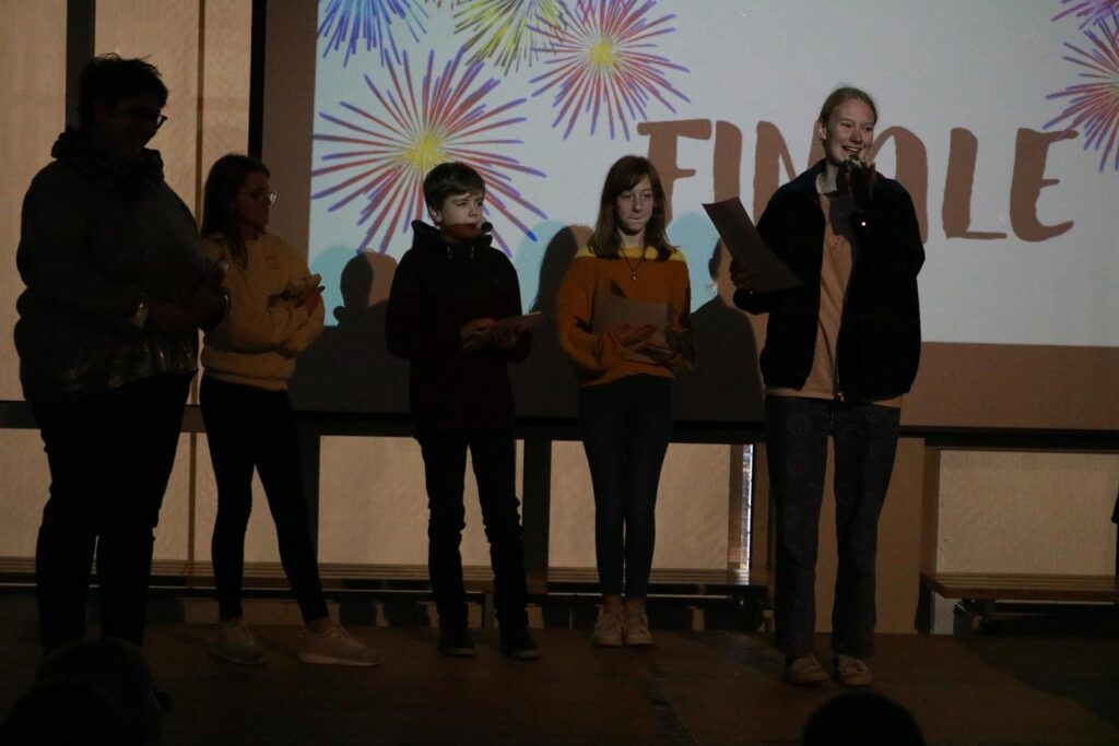 Schüler präsentieren auf einer Bühne ein Finale mit bunten Feuerwerk-Visuals im Hintergrund.