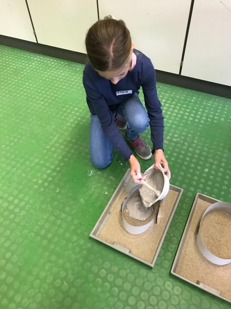 Mädchen schüttet Sand aus einem Behälter in zwei Formen auf einem grünen Boden.
