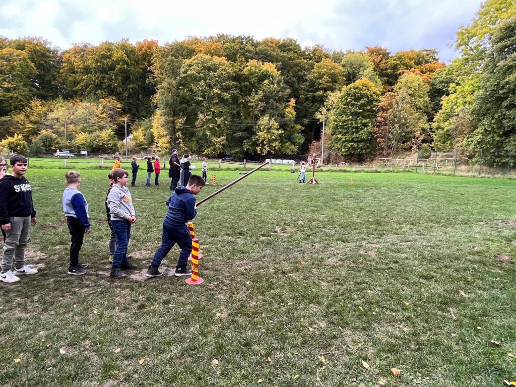 Kinder stehen in einer Reihe und üben Wurfspiel im Freien. Bäume im Hintergrund.