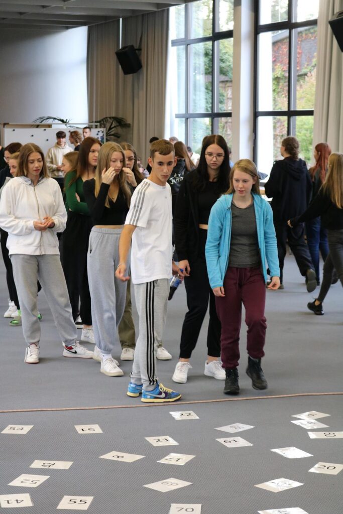 Schüler bewegen sich in einer Aula zwischen auf dem Boden liegenden Zetteln.