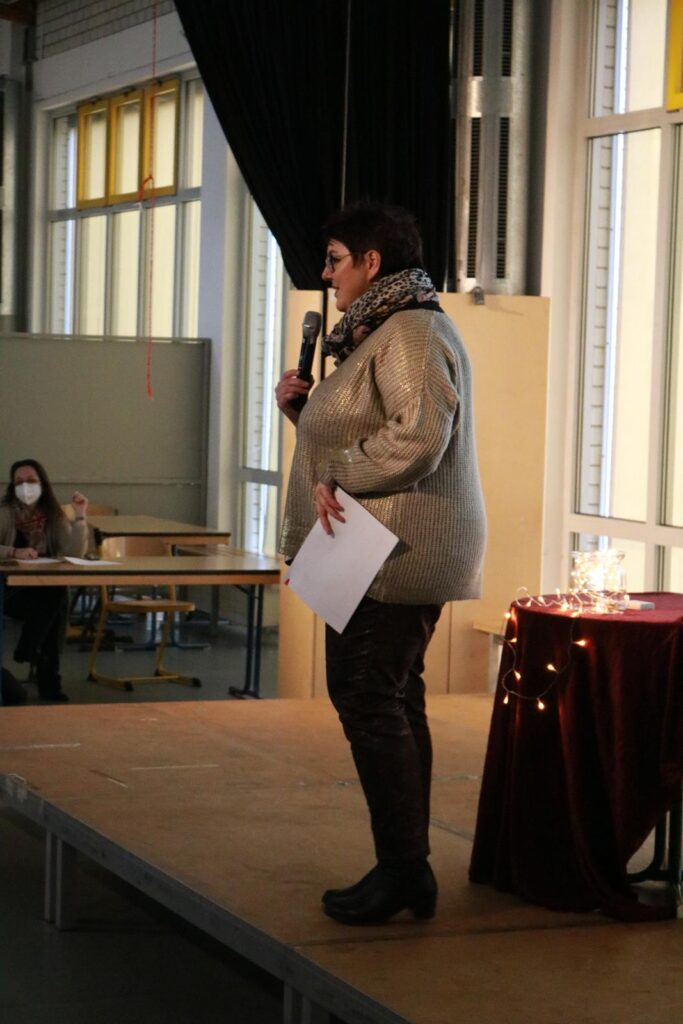 Frau spricht auf einer Bühne vor einem Publikum in einem Schulraum.