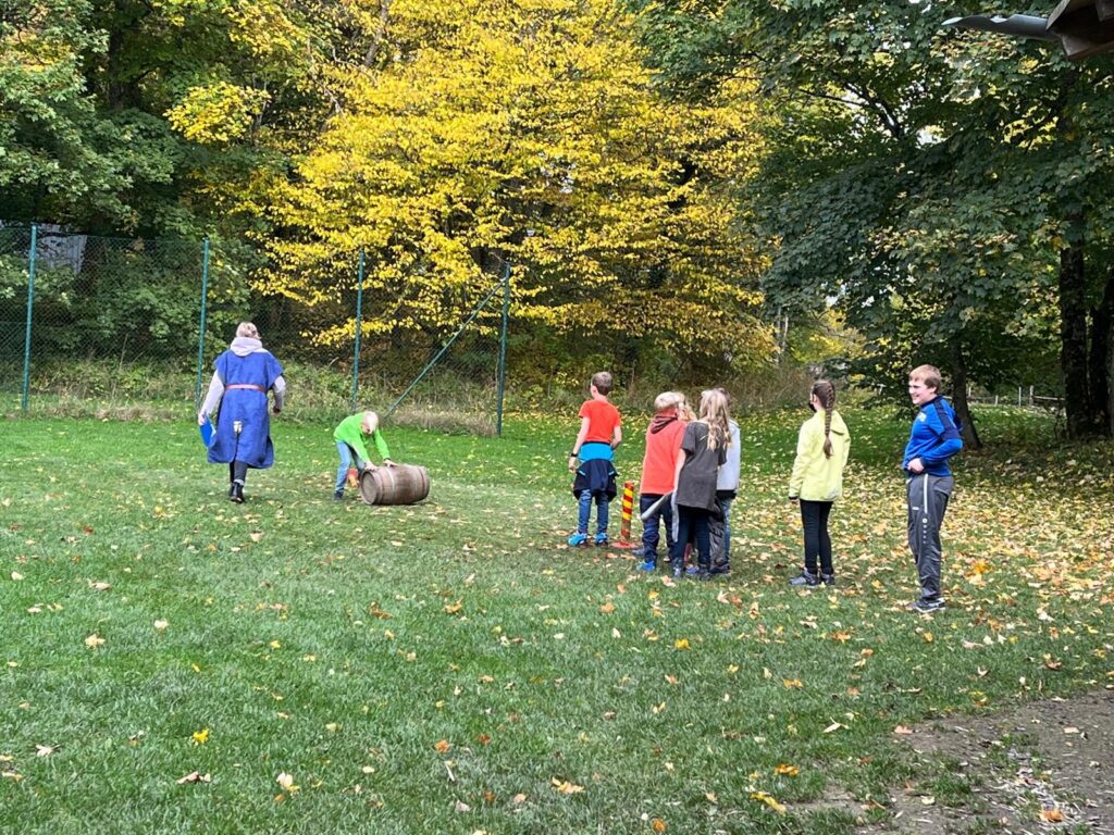Kinder spielen im Freien, eine Person rollt eine große Rolle. Bäume im Hintergrund.