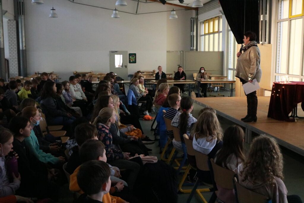 Schülerin spricht vor Publikum in einer Aula mit Stühlen und Tischen.