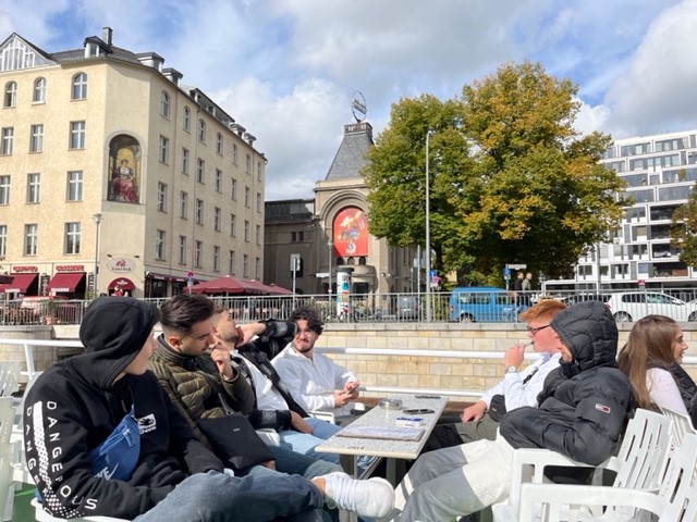 Gruppe junger Menschen sitzt an einem Tisch im Freien, Stadtansicht im Hintergrund.