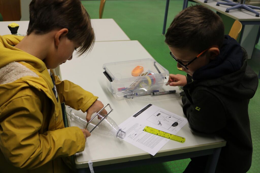Zwei Kinder arbeiten an einem Tisch mit Materialien und Anweisungen für ein Experiment.