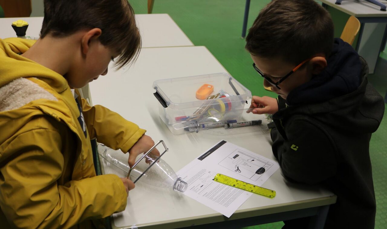 Zwei Kinder arbeiten an einem Tisch mit Materialien und Anweisungen für ein Experiment.