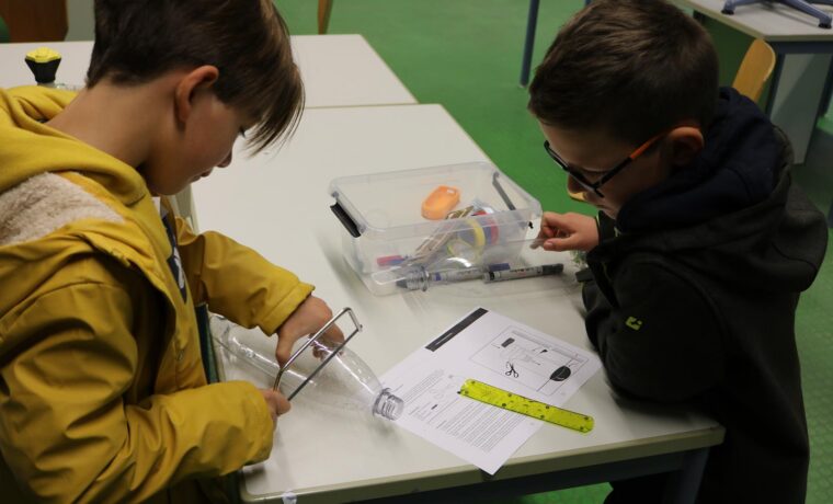 Zwei Kinder arbeiten an einem Tisch mit Materialien und Anweisungen für ein Experiment.