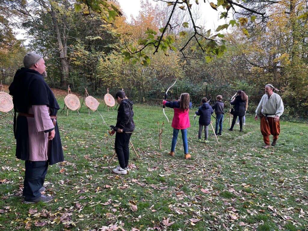 Kinder üben mit Bögen im Freien, Erwachsene beobachten und unterstützen. Herbstliche Umgebung.