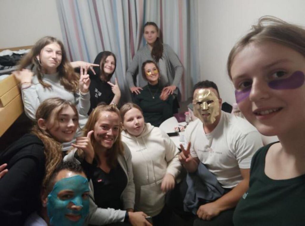 Gruppenselfie mit Mädchen und einem Jungen, alle mit Gesichtsmasken, in einem Zimmer.