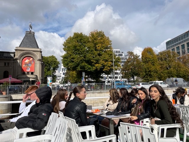 Schüler sitzen an einem Tisch im Freien, im Hintergrund Bäume und Gebäude.