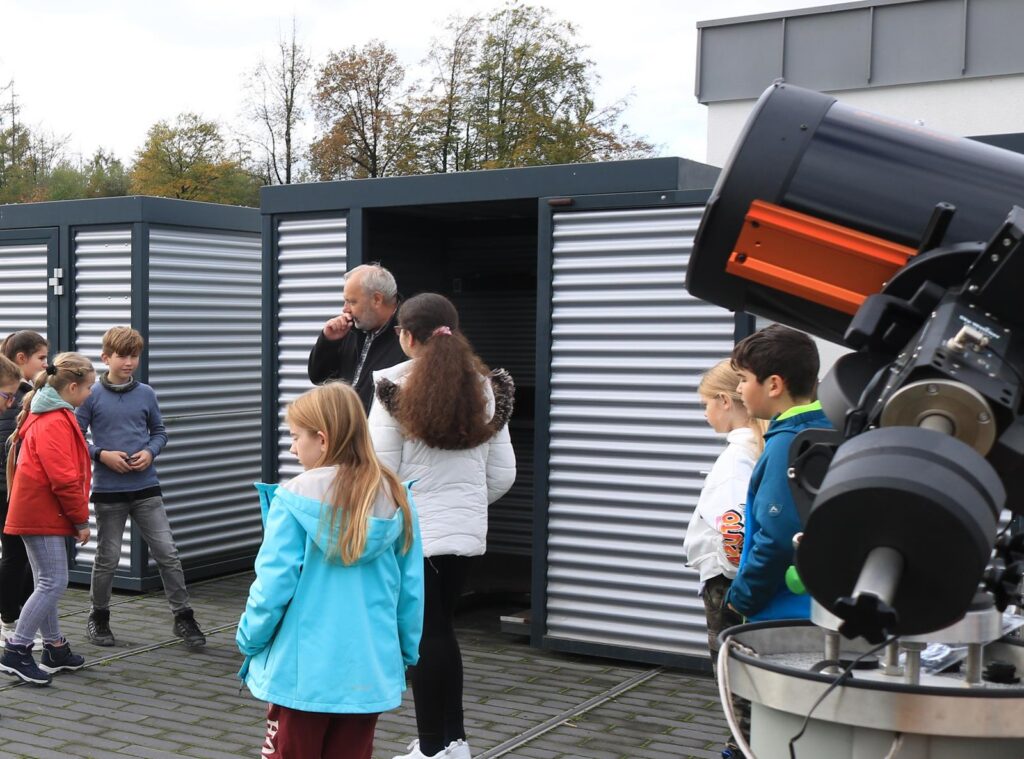 Gruppe von Kindern und Erwachsenen vor observatoriumartigen Gebäuden mit einem Teleskop.
