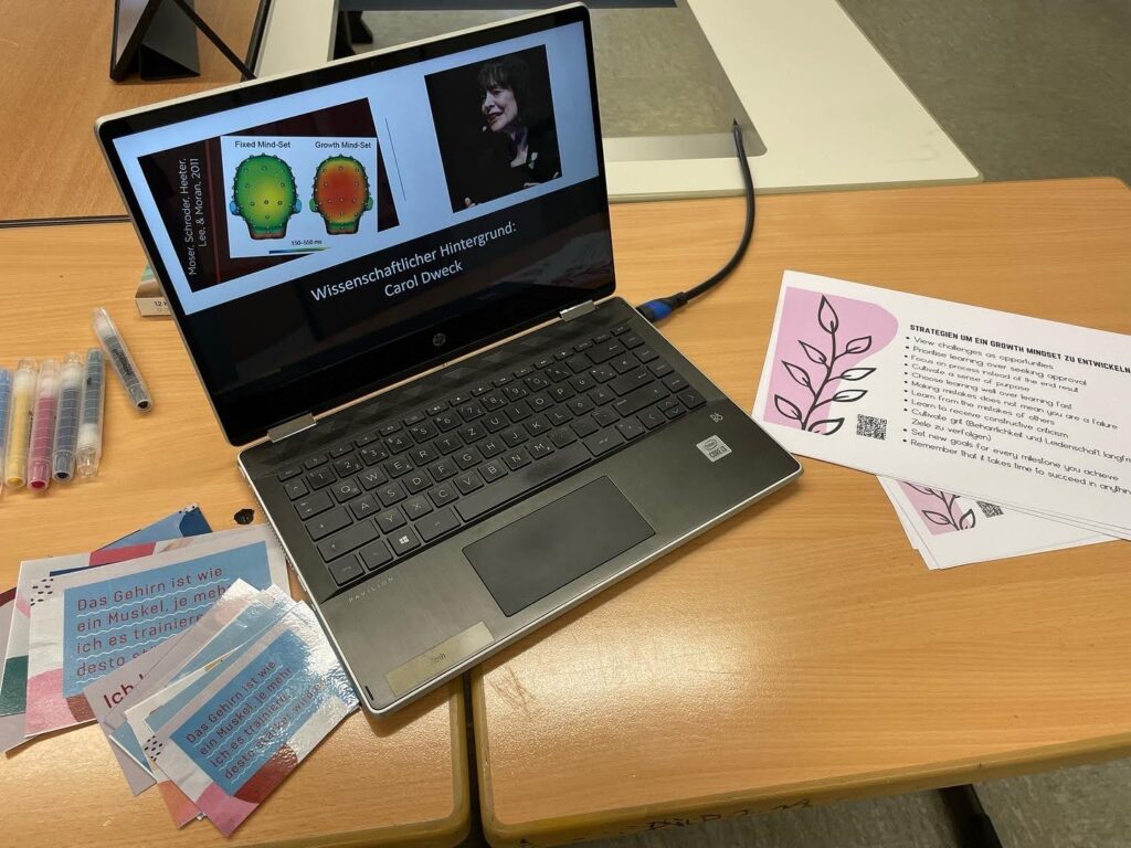 Laptop mit Präsentation und Materialien auf einem Tisch. Schreibutensilien daneben.