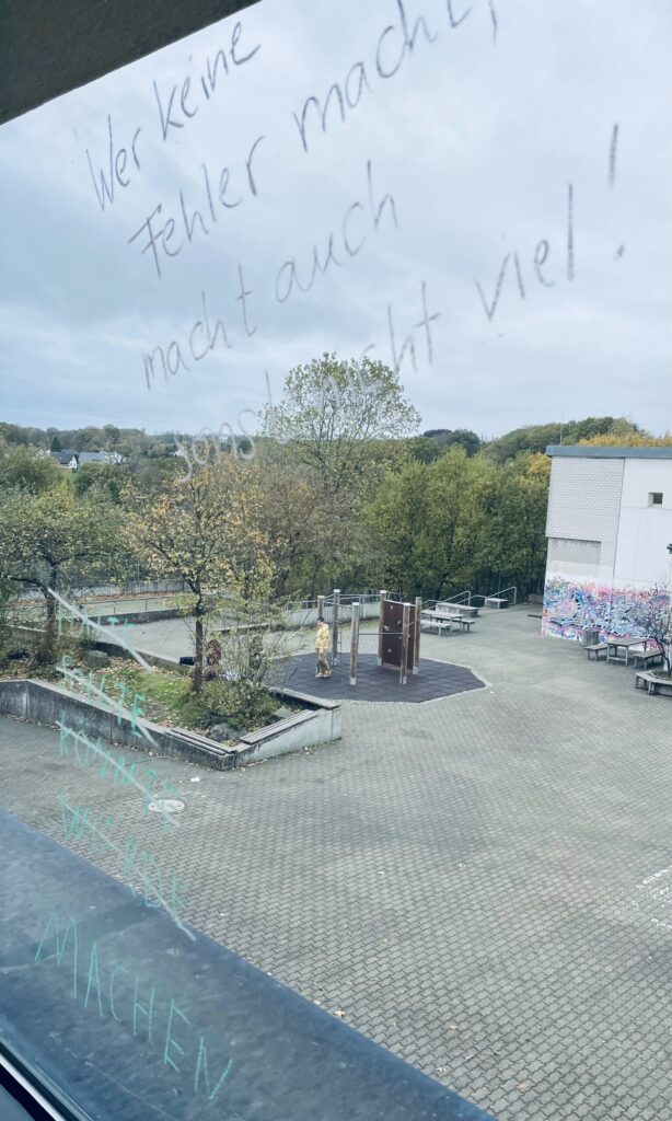 Ausblick auf einen Schulhof mit grauem Himmel und handgeschriebener Nachricht am Fenster.