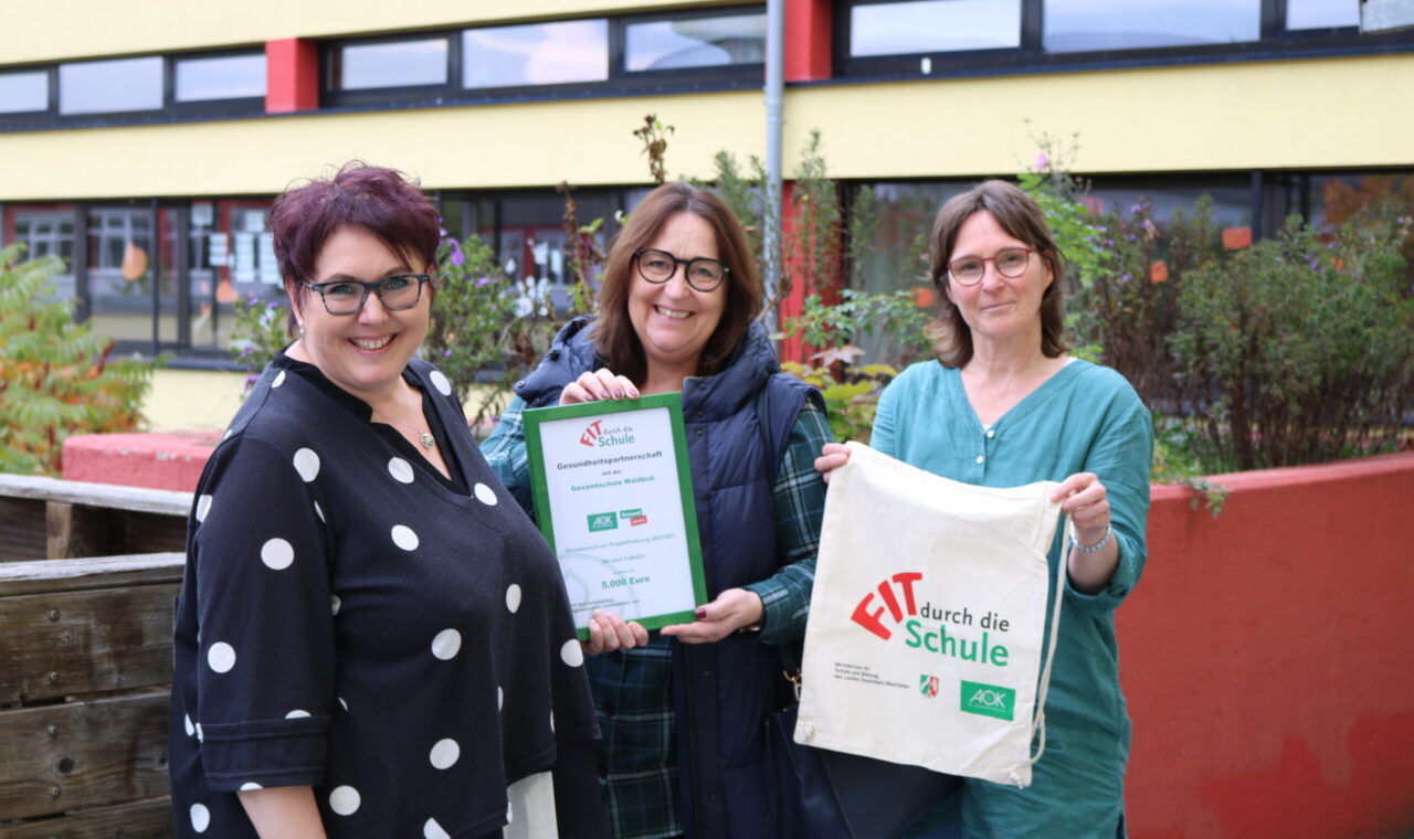 Drei Frauen halten eine Urkunde und eine Tasche vor einer Schulgebäude.