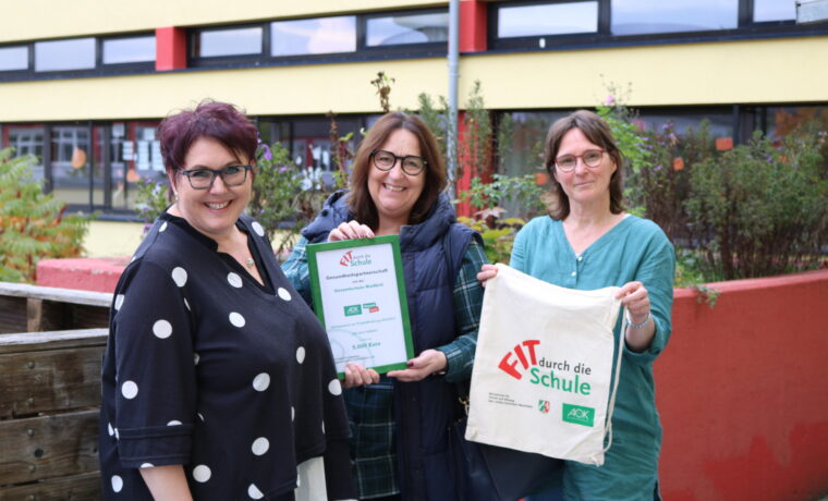 Drei Frauen halten eine Urkunde und eine Tasche vor einer Schulgebäude.