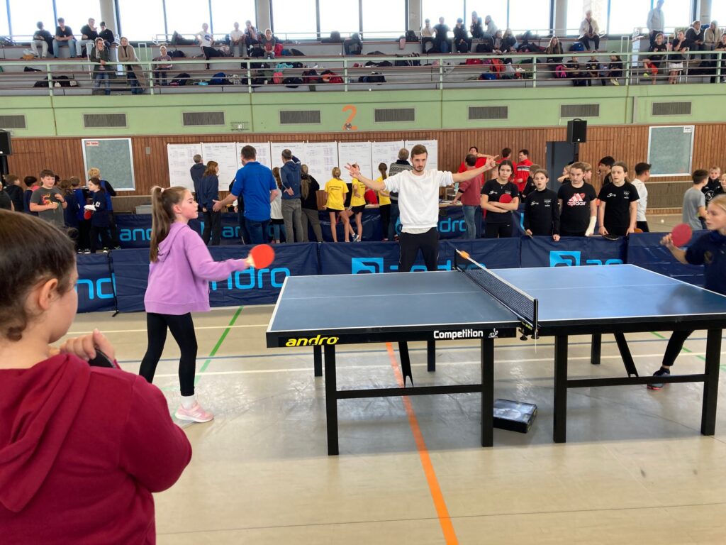 Tischtennisturnier mit Spielern, Zuschauern und mehreren Tischen in einer Sporthalle.
