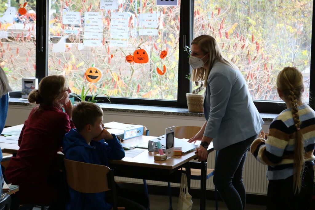 Lehrerin spricht mit Schülern in einem Klassenraum. Herbstdekoration sichtbar.