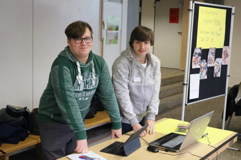 Zwei Schüler stehen an einem Tisch mit Laptops und Präsentationsmaterialien.