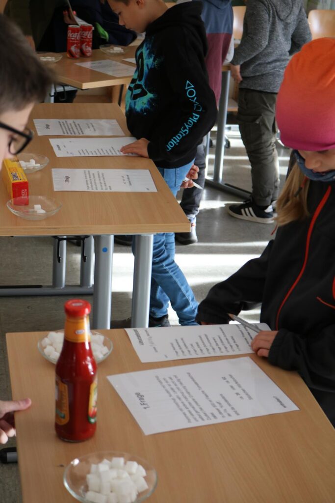 Schüler arbeiten an Tischen mit Arbeitsblättern und Materialien wie Zuckerwürfeln und Flaschen.