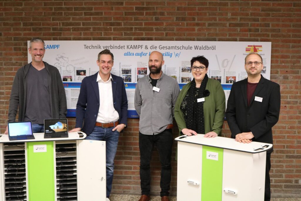 Personengruppe steht an Tischen mit Technik-Geräten vor einer Wand.