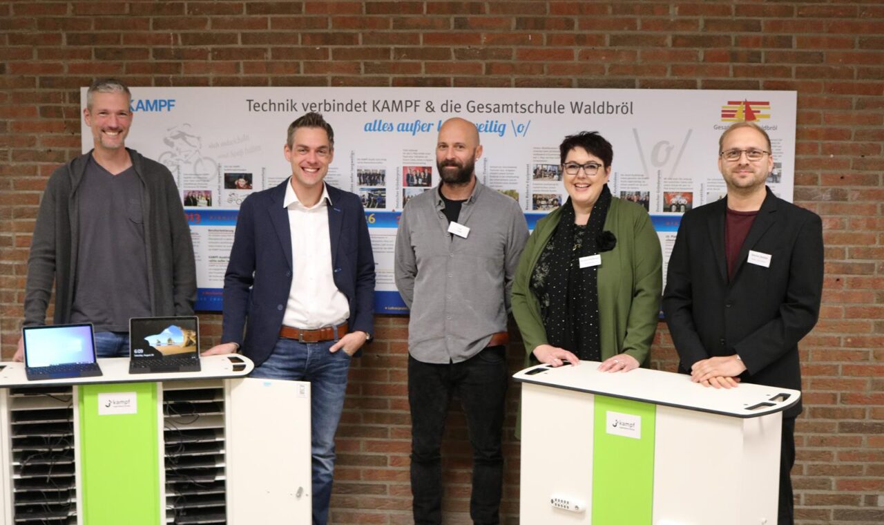 Personengruppe steht an Tischen mit Technik-Geräten vor einer Wand.