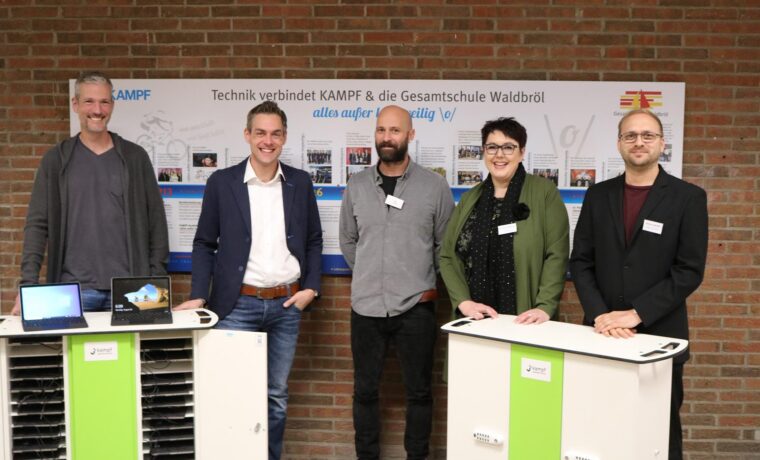 Personengruppe steht an Tischen mit Technik-Geräten vor einer Wand.
